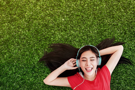 快乐的年轻女子躺在绿草上，通过耳机在公园里听音乐。 微笑着闭上眼睛俯视