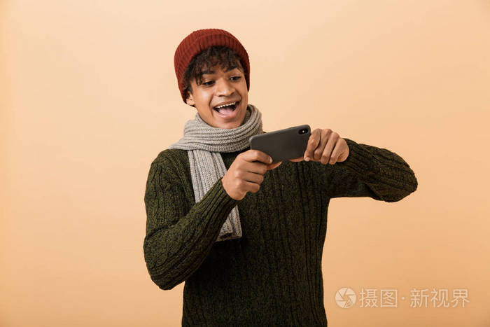 快乐的年轻非洲男人的肖像穿着秋装被隔离在米色背景下在手机上玩游戏