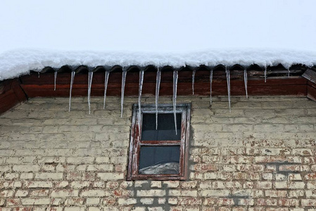 屋顶上的一排长长的冰柱，雪下的一座棕色砖墙上有一扇窗户
