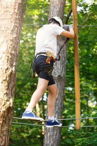 森林探险公园的孩子。 穿着橙色头盔和蓝色T恤的孩子爬上高绳小径。 敏捷技能和攀爬儿童户外娱乐中心。 小男孩在户外玩耍