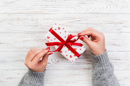 女人的手在纸上用红色丝带包裹情人节礼物或其他节日手工礼物。礼物盒，红心装饰礼品在木桌上，顶部的视野与复制空间为您设计。