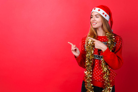 一个穿着红色毛衣，戴着脖子上戴着tinsel的圣诞老人帽子的漂亮女孩在红色背景上的一个空地上展示了她的手指。图片