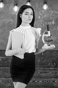 夫人科学家持有书籍和显微镜, 黑板上的背景, 复制空间。女士在正式穿戴在安静的面孔在教室。生物学老师拥有书本和显微镜。生物学概念