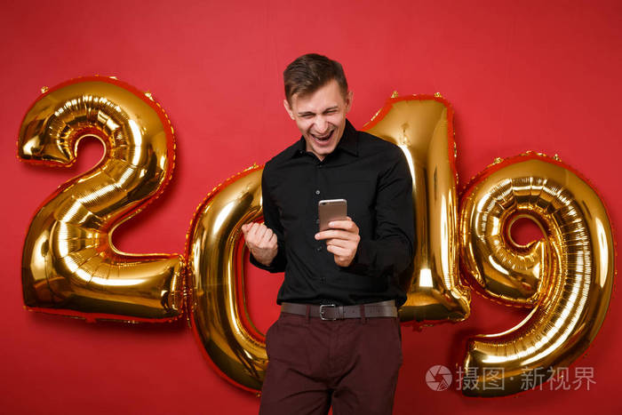 快乐有趣的年轻人穿着黑色衬衫庆祝节日聚会，手持手机隔离在明亮的红色墙壁背景，金色数字气球工作室的肖像。新年快乐2019圣诞概念