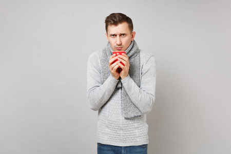 冰冻的年轻人穿着灰色毛衣围巾，拿着红色的咖啡或茶，孤立在灰色背景上，在工作室。 健康生活方式病态疾病治疗寒冷季节的概念。 模拟复