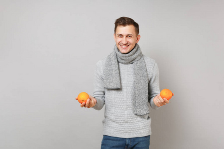 微笑的年轻人戴着灰色毛衣围巾，抱着灰色墙壁背景工作室肖像上孤立的橘子。 健康时尚生活方式人真诚情感冷季观念。 模拟复制空间