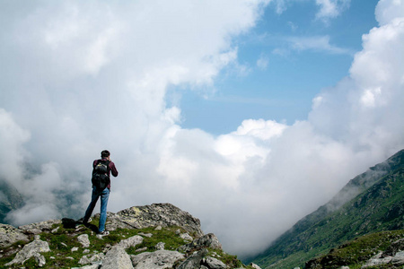 游客背着一个大背包，站在山顶，手里拿着一个动作摄像机，拍照