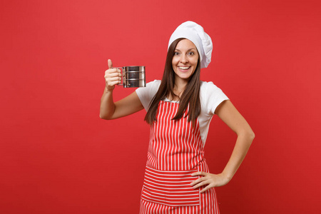家庭主妇女厨师或面包师穿条纹围裙，白色T恤，烤厨师帽子隔离在红墙背景上。 管家女人拿着金属铁筛筛筛面粉。 模拟复制空间概念