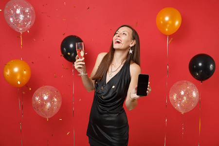 快乐的女人穿着黑色的裙子抬头，拿着手机，空白的黑色屏幕香槟在明亮的红色背景气球上。新年快乐，生日模拟节日聚会理念