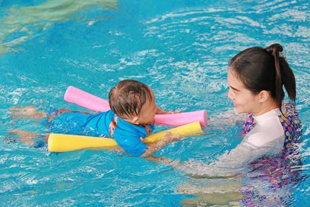 年轻的亚洲妈妈在游泳池里用面条泡沫教小男孩。