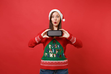 惊讶的年轻圣诞老人女孩保持嘴巴张开，看着惊讶地拿着便携式无线蓝牙音乐扬声器隔离在红色背景上。 新年快乐2019庆祝节日派对概念