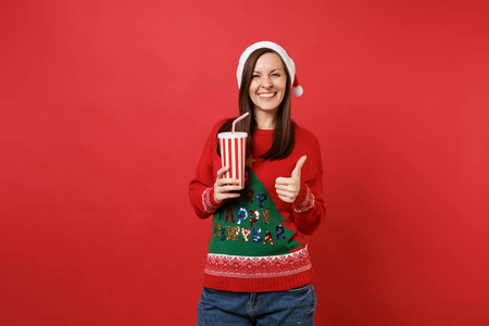 微笑的年轻圣诞老人女孩戴着圣诞帽，拇指向上举着塑料杯汽水或可乐，孤立在红色背景上。 新年快乐，2019年庆祝节日聚会的理念。 模