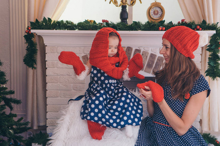 一个女人带着一个穿着蓝色连衣裙的孩子，戴着红色帽子和手套的圆点，背景是圣诞节内部壁炉圣诞树礼物手表