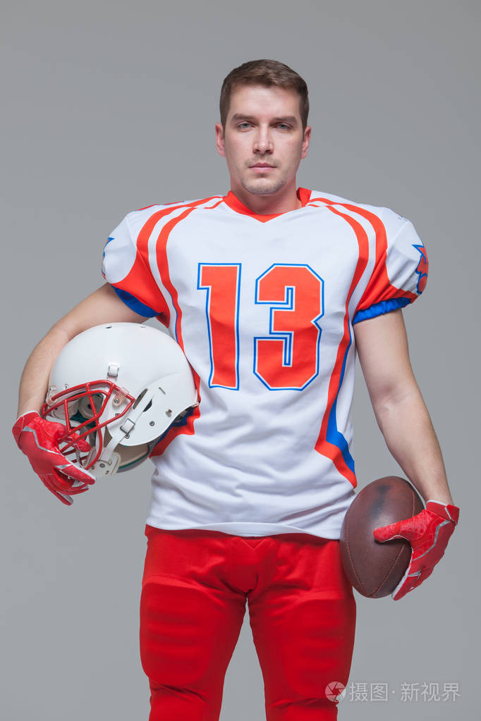 美国足球运动员拿着橄榄球球在灰色背景的正面观下戴头盔