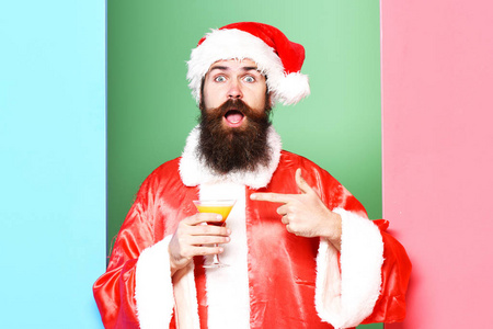 英俊的胡须圣诞老人男子与长胡子惊讶的脸上拿着一杯酒精饮料在圣诞节或圣诞毛衣和新年帽子显示在五颜六色的工作室背景