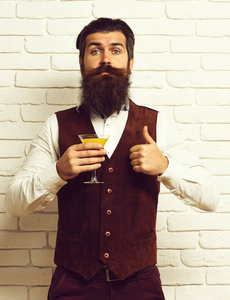 英俊的大胡子的男人，留着长胡子，胡子含酒精的饮料杯举行复古绒面皮革背心白砖壁演播室背景上的滑稽脸上有时尚的发型