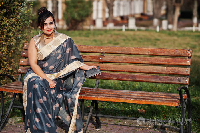 优雅的黑发南亚印度女孩在萨雷坐在长凳上手里拿着手机