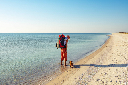 在一个阳光明媚的日子里，带着有趣的狗的旅行者沿着绿松石海旁边的荒凉海滩走去。 可怕的冒险旅行到海滨。 后景。