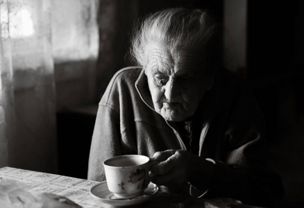 黑白影像的一个老抑郁的女人..一位年迈的孤独女子坐在靠近窗户的厨房的一张桌子旁喝茶。