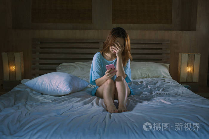 年轻抑郁的亚洲韩国女孩使用手机在床上哭泣,晚上感到悲伤和沮丧,网络