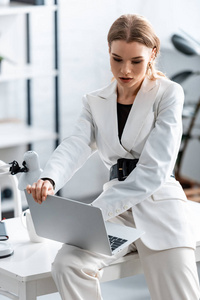 穿着白色正装坐在办公桌上在工作场所使用笔记本电脑的漂亮专注的女商人
