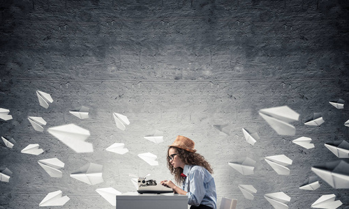 年轻而美丽的女作家戴着帽子和眼镜，使用打字机，坐在飞机上的桌子上，靠在背景上灰色的混凝土墙上。
