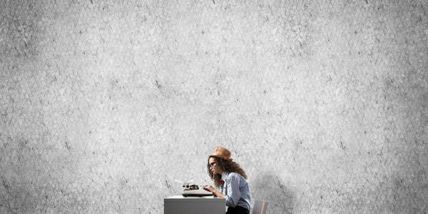 年轻漂亮的女作家戴着帽子和眼镜，一边坐在桌子旁，背景上是灰色的混凝土墙。