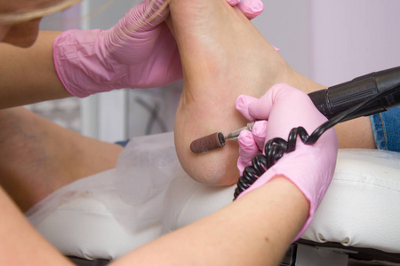 专业五金修脚使用粉红色手套和电动装置机。 病人在医学修脚术程序访问足科医生。 剥皮的脚。 水疗中心脚部治疗。脚部诊所