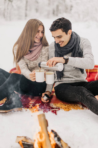 一对年轻夫妇在冬天的森林里约会，被火加热，喝可可