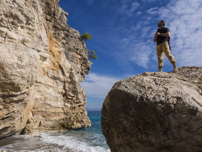 带背包的旅行者在靠近大海的岩石上望去。暑期旅游假期。英俊的年轻的白种人游客在户外休闲服装自然