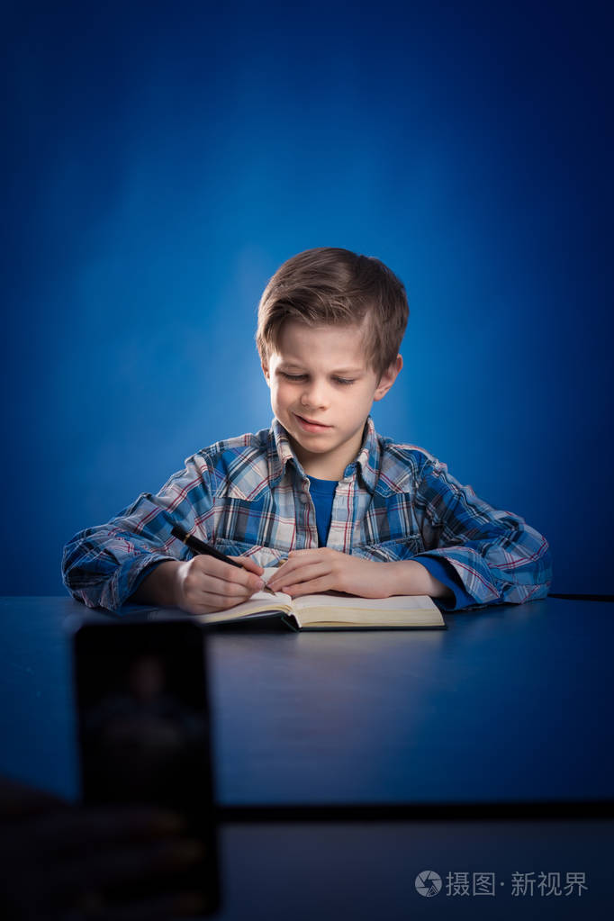 一个穿格子衬衫的小男孩坐在桌子旁,在蓝色背景上的笔记本上写字