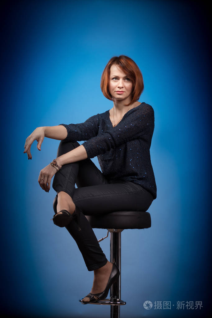 情绪激动的红发女人,穿着黑色衣服坐在酒吧凳子上,在蓝色背景上摆姿势