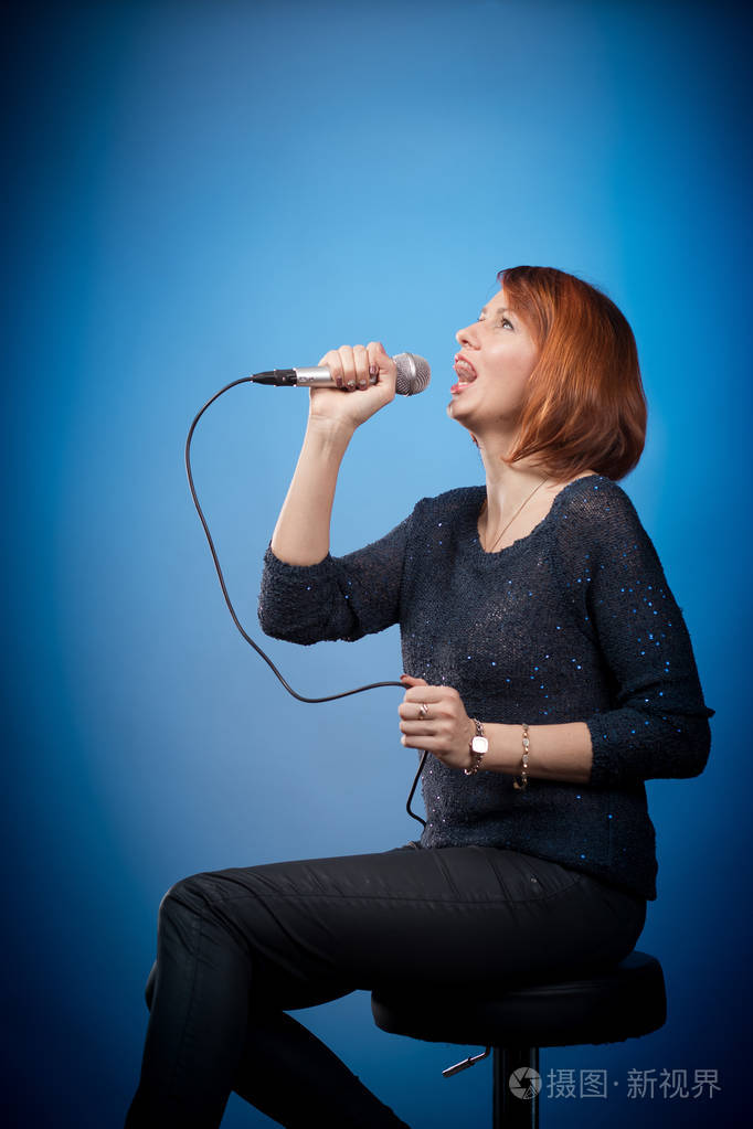 红发女人穿着黑色衣服,拿着麦克风坐在酒吧凳子上,在蓝色背景上唱歌