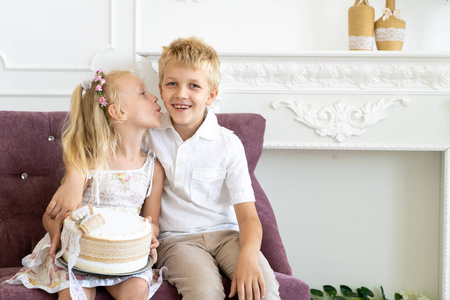 可爱的小男孩和漂亮的女孩坐在椅子上。 妹妹祝贺他哥哥生日。 拿着一个大的白色蛋糕和洒满奶油。 干净衬衫上衣