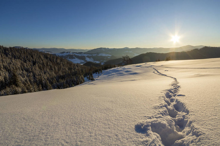 冬季圣诞景观。 人类足迹轨迹在水晶白色的深雪中穿过空旷的田野，木质的黑暗山脉，在清澈的蓝天上柔和地发光，复制空间背景。