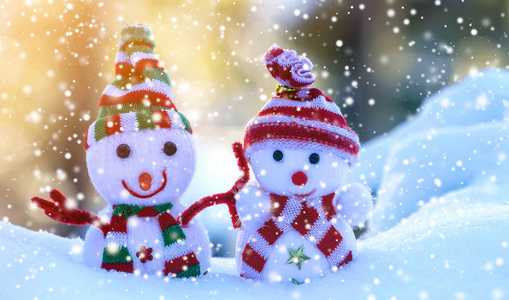 两个小有趣的玩具婴儿雪人在针织帽子和围巾在深雪户外明亮的蓝色和白色复制空间背景。 新年快乐，圣诞贺卡快乐。