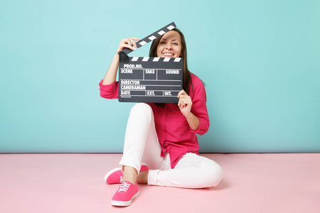 全长镜头的女人，穿着玫瑰衬衫，白色裤子，坐在地板上，与电影制作剪贴板隔离在粉红色的蓝色粘贴墙背景工作室。 时尚生活方式概念模拟复