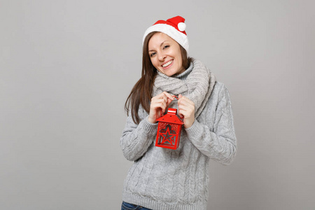 漂亮的年轻圣诞老人女孩戴着灰色毛衣围巾，戴着圣诞帽，手持老式灯笼烛台，灰色背景。 新年快乐，2019年庆祝节日聚会的理念。 模拟