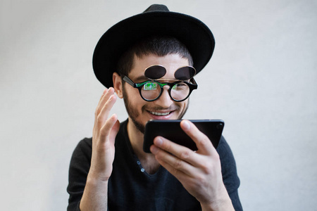 快乐的小胡子嬉皮士在白色背景下玩智能手机游戏。戴着眼镜和帽子的黑色衣服