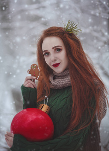 红头发的长发微笑的女孩与一个巨大的圣诞红球吃姜饼。一个穿着毛衣的红头发的年轻女子走在雪堆里