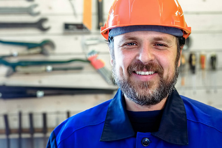 一个戴着头盔和工作服的胡须的人在一个带工具的支架的背景下微笑着雪白的微笑。