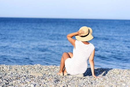 年轻的黑发女人在夏天白色礼服座位上的海滩上, 并期待大海。白种人女孩放松和享受假期的和平
