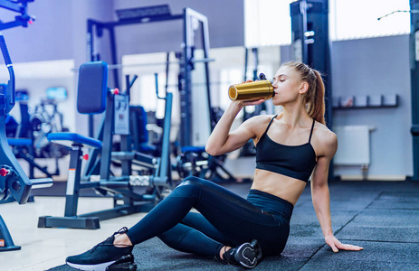 健身妇女在健身房里坐在地板上休息时喝水。 妇女在健身健身房锻炼，打破放松，并在训练期间持有蛋白摇瓶。