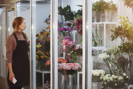 在花卉沙龙穿棕色围裙的女花店接受在线订购。 选定的焦点模糊背景。