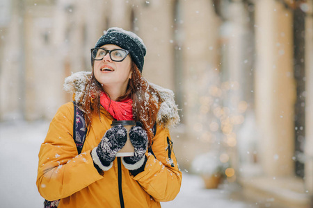 美丽的年轻女孩喝咖啡在雪的冬天的城市环顾四周