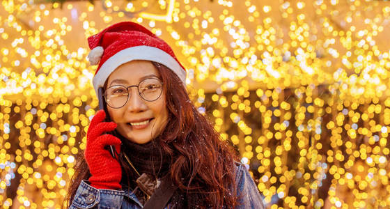 一位美丽快乐的亚洲女孩戴着一顶红色圣诞老人帽子，戴着一部智能手机，站在灯光和圣诞花环附近，在冬天下雪时微笑着。