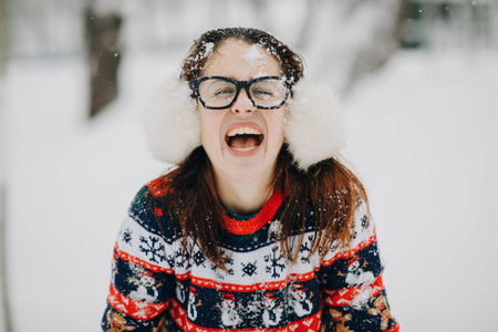 户外近距离冬季肖像的年轻美丽的女孩戴耳罩, 毛衣摆在雪公园。看照相机和微笑的妇女