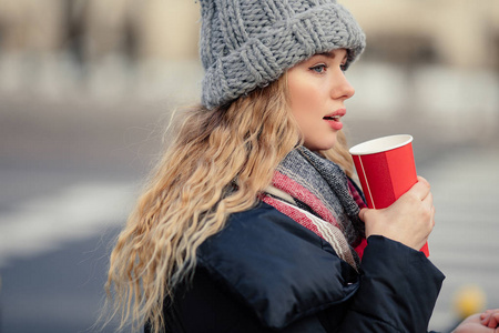 女人在街上散步时喝她的热咖啡。 穿着冬天衣服喝热咖啡的时尚微笑女人的肖像。 女性冬季风格。 图像