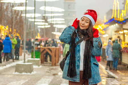 一个戴着红色圣诞老人帽子的美丽的亚洲女孩站在城市的灯光和圣诞灯附近，在冬天里带着雪微笑。