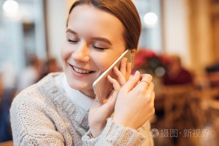 快乐的女孩坐在咖啡馆里，用快乐的脸在电话里交谈。 穿白色毛衣。 冬季秋装。